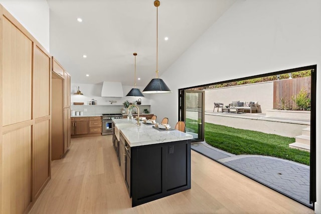 kitchen with light stone counters, wall chimney exhaust hood, a kitchen island with sink, stainless steel stove, and hanging light fixtures