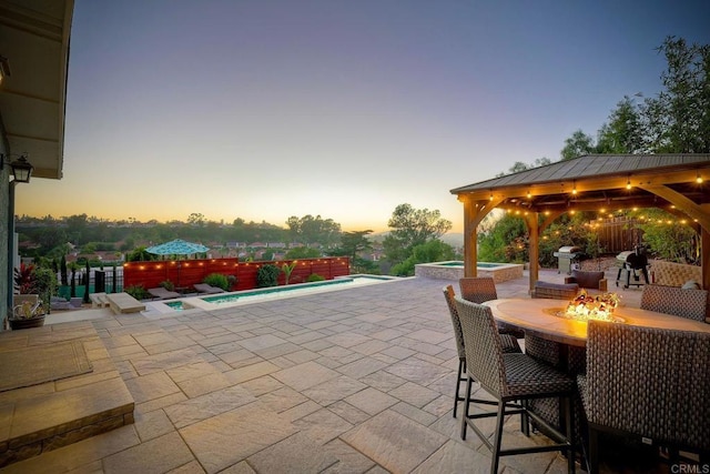 patio terrace at dusk with a gazebo, grilling area, a fire pit, and a fenced in pool