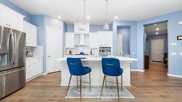 kitchen with appliances with stainless steel finishes, wall chimney exhaust hood, decorative light fixtures, white cabinets, and a kitchen island with sink