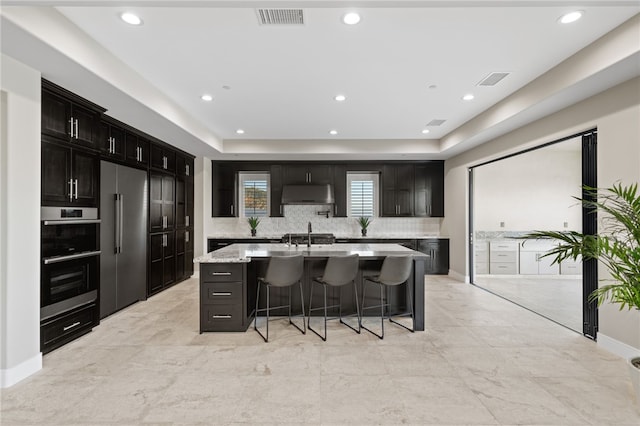 kitchen featuring a kitchen breakfast bar, tasteful backsplash, light stone counters, a center island with sink, and appliances with stainless steel finishes