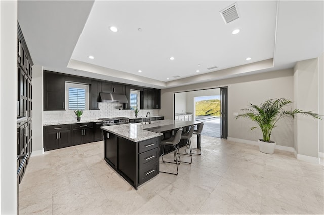 kitchen featuring stainless steel stove, a raised ceiling, a kitchen island with sink, and a breakfast bar area