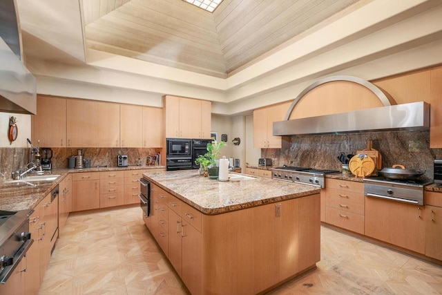 kitchen with range hood, a center island, a tray ceiling, light brown cabinetry, and light stone counters