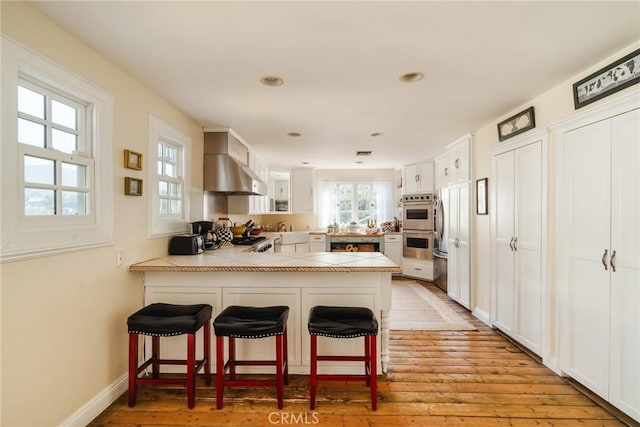 kitchen featuring white cabinets, a kitchen breakfast bar, wall chimney range hood, light wood-type flooring, and kitchen peninsula