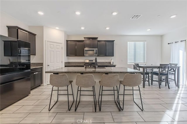 kitchen with dark stone counters, dark brown cabinets, stainless steel appliances, sink, and an island with sink