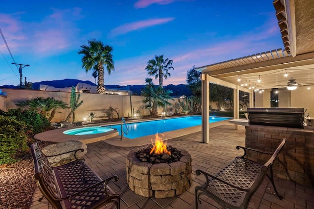 pool at dusk featuring an in ground hot tub, a mountain view, a fire pit, area for grilling, and a patio area