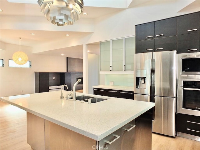 kitchen featuring appliances with stainless steel finishes, an island with sink, sink, hanging light fixtures, and light hardwood / wood-style flooring