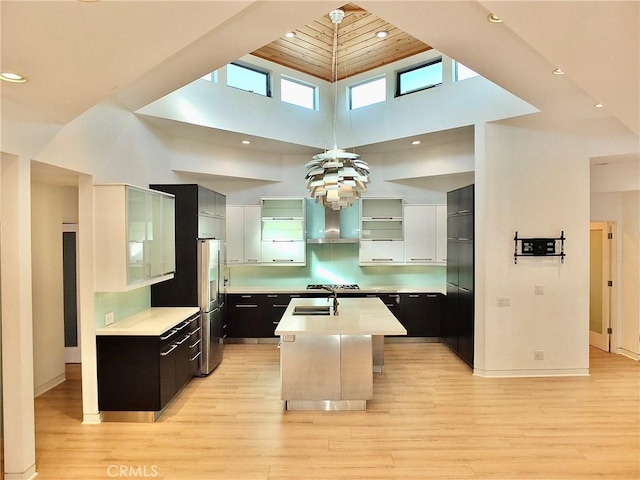 kitchen featuring wall chimney range hood, light hardwood / wood-style floors, a high ceiling, and a center island with sink