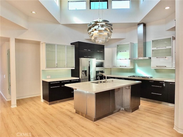 kitchen with sink, a towering ceiling, stainless steel appliances, an island with sink, and wall chimney exhaust hood