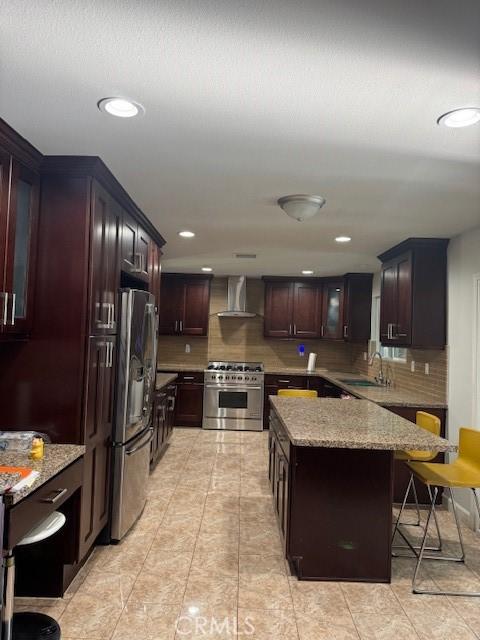 kitchen with backsplash, a kitchen breakfast bar, wall chimney range hood, appliances with stainless steel finishes, and light stone counters