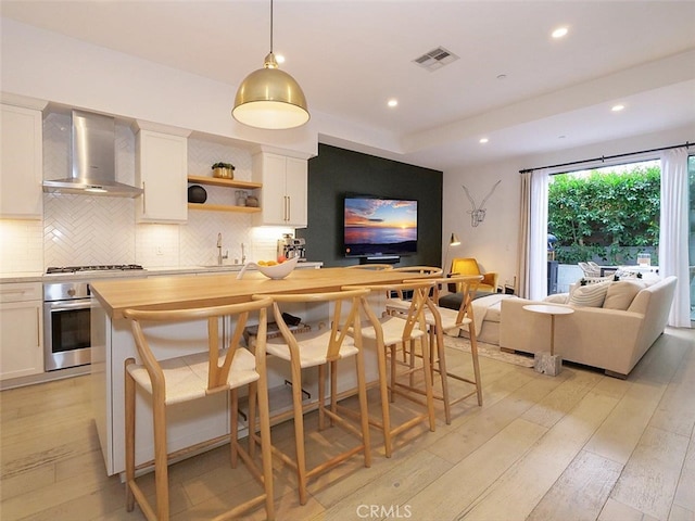 kitchen featuring wall chimney range hood, wooden counters, white cabinets, a kitchen bar, and stainless steel oven