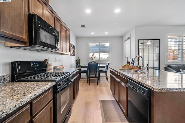 kitchen with light stone countertops, light wood-type flooring, sink, black appliances, and a center island with sink