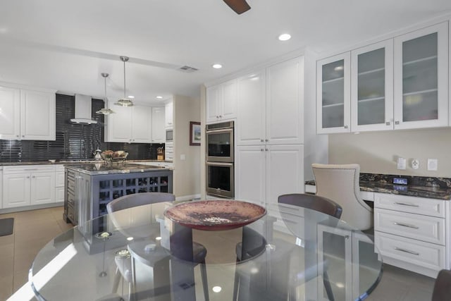 kitchen featuring decorative light fixtures, stainless steel double oven, wall chimney range hood, and white cabinetry