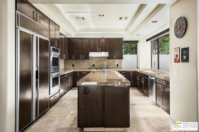 kitchen with stainless steel appliances, an island with sink, dark brown cabinetry, a tray ceiling, and sink