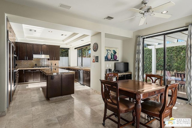 kitchen featuring a center island with sink, ceiling fan, a tray ceiling, backsplash, and dark brown cabinetry