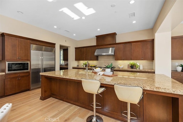 kitchen featuring built in appliances, an island with sink, a breakfast bar area, light stone countertops, and range hood