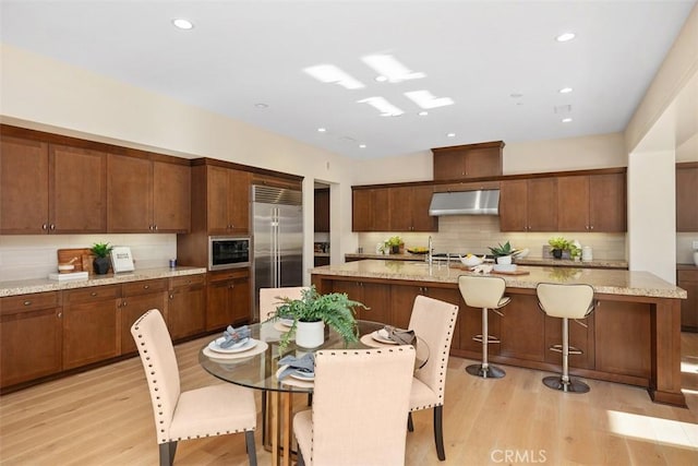 kitchen featuring light wood-style floors, backsplash, built in appliances, under cabinet range hood, and a sink