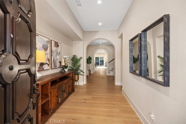 hallway featuring arched walkways, light wood finished floors, recessed lighting, baseboards, and stairs