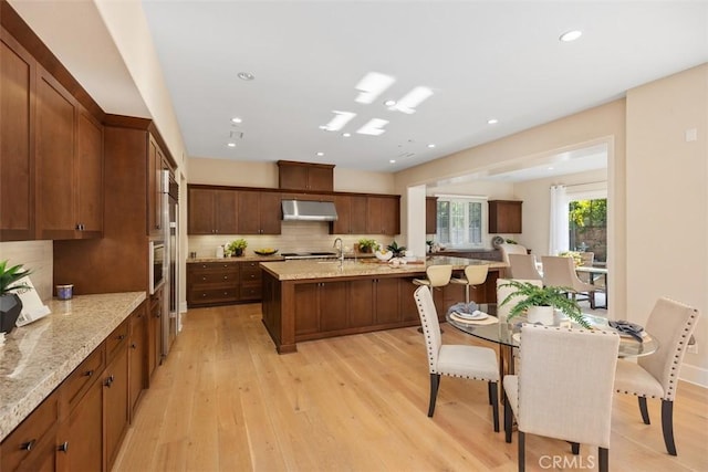 kitchen with ventilation hood, an island with sink, light wood-type flooring, and stainless steel oven