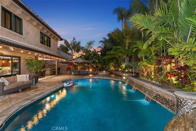 pool at dusk featuring outdoor lounge area, an outdoor kitchen, pool water feature, and a patio