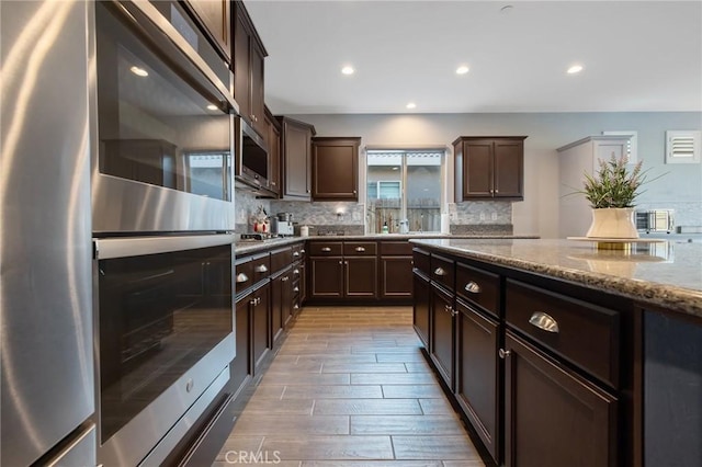 kitchen with stainless steel appliances, decorative backsplash, dark brown cabinets, and light stone countertops