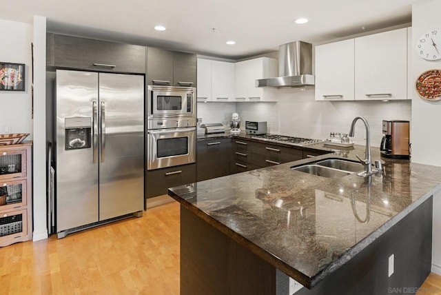 kitchen featuring light hardwood / wood-style flooring, wall chimney range hood, dark stone counters, appliances with stainless steel finishes, and sink