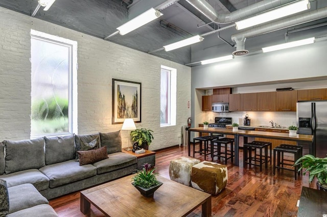 living room featuring a towering ceiling, brick wall, dark wood-type flooring, and plenty of natural light