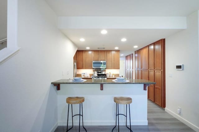 kitchen with light hardwood / wood-style floors, kitchen peninsula, appliances with stainless steel finishes, a kitchen breakfast bar, and dark stone counters