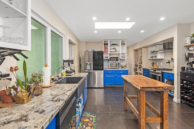 kitchen with light stone counters, wall chimney exhaust hood, a skylight, blue cabinets, and appliances with stainless steel finishes