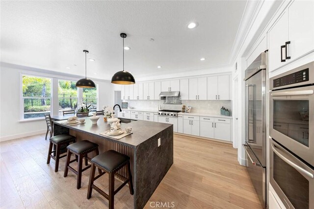 kitchen featuring stainless steel appliances, white cabinets, hanging light fixtures, tasteful backsplash, and a center island with sink