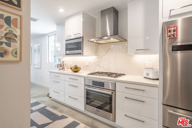 kitchen with light stone countertops, stainless steel appliances, wall chimney range hood, tasteful backsplash, and white cabinetry