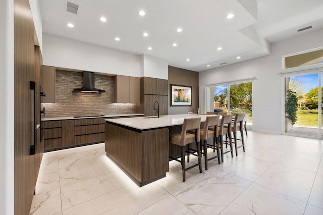 kitchen featuring a kitchen island with sink, black cooktop, a kitchen bar, wall chimney exhaust hood, and sink