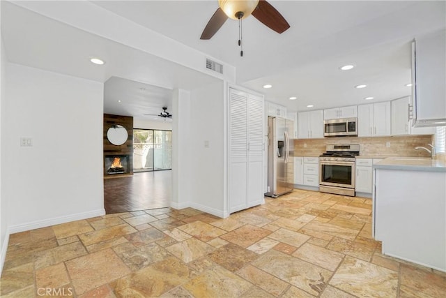 kitchen featuring appliances with stainless steel finishes, ceiling fan, decorative backsplash, a fireplace, and white cabinetry