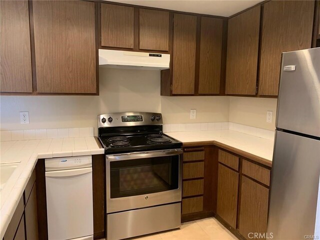 kitchen featuring tile counters, appliances with stainless steel finishes, light tile patterned floors, and dark brown cabinets