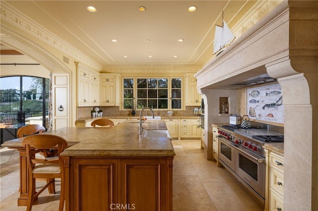 kitchen featuring double oven range, decorative backsplash, a kitchen island with sink, cream cabinetry, and sink