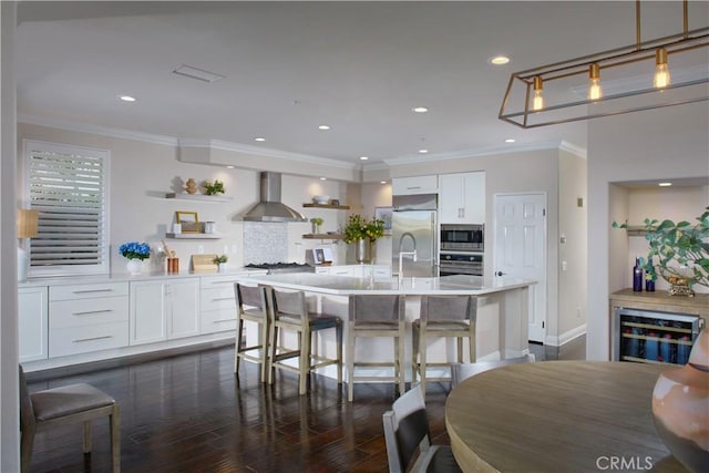 kitchen with built in appliances, an island with sink, wall chimney exhaust hood, and white cabinetry