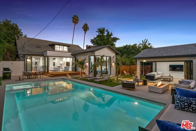 pool at dusk with a deck, a patio, an outbuilding, grilling area, and an outdoor living space with a fire pit