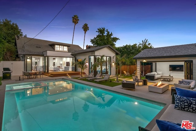 pool at dusk with a deck, a patio, an outbuilding, grilling area, and an outdoor living space with a fire pit