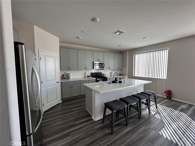 kitchen with dark wood-type flooring, gray cabinetry, appliances with stainless steel finishes, an island with sink, and decorative backsplash