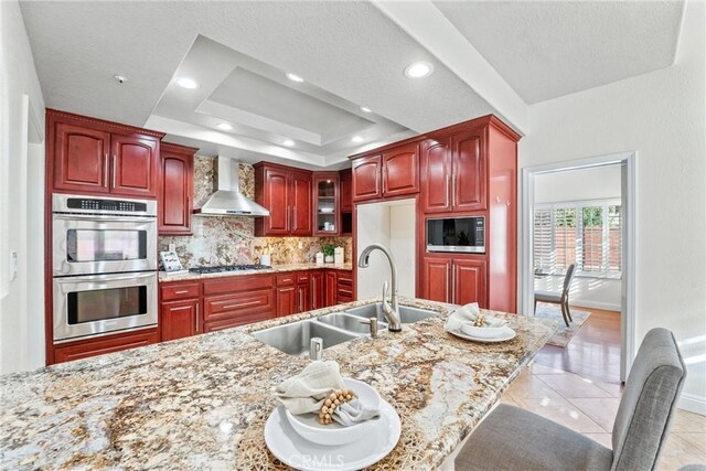 kitchen with sink, appliances with stainless steel finishes, a tray ceiling, light stone countertops, and wall chimney exhaust hood