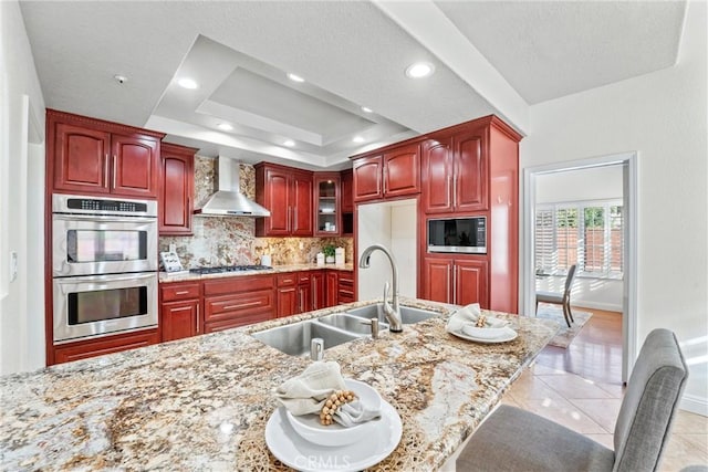 kitchen with stainless steel appliances, light stone counters, wall chimney range hood, a tray ceiling, and sink