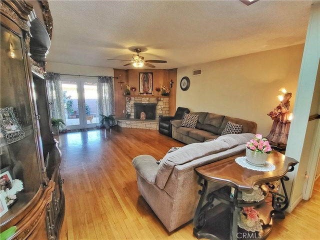 living room featuring a textured ceiling, light hardwood / wood-style flooring, french doors, ceiling fan, and a stone fireplace