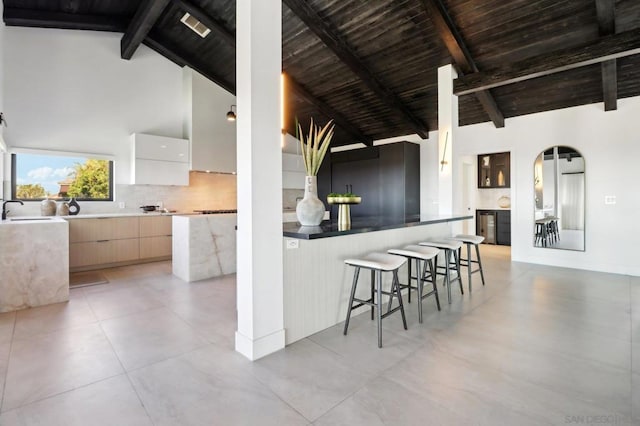 kitchen featuring a breakfast bar area, high vaulted ceiling, decorative backsplash, beam ceiling, and white cabinets