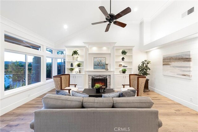 living room featuring light wood-type flooring, ceiling fan, crown molding, and a stone fireplace