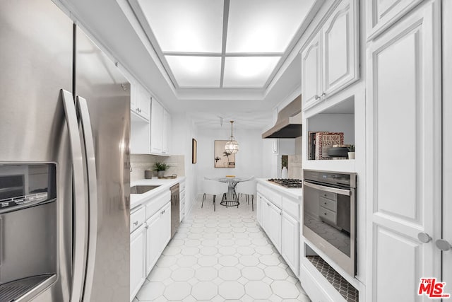 kitchen with wall chimney range hood, white cabinetry, backsplash, stainless steel appliances, and decorative light fixtures