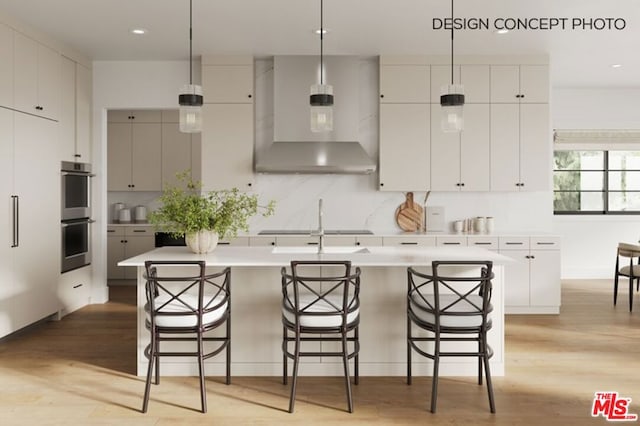 kitchen with stainless steel double oven, an island with sink, wall chimney range hood, and pendant lighting