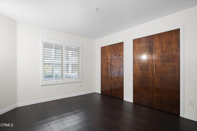 unfurnished bedroom with baseboards, dark wood-type flooring, and two closets