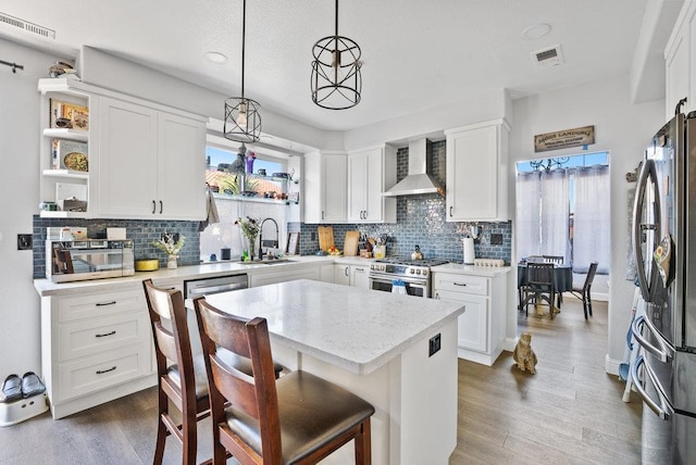 kitchen featuring white cabinets, a kitchen island, wall chimney range hood, stainless steel appliances, and hanging light fixtures