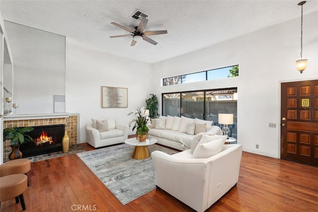 living room featuring a tile fireplace, ceiling fan, hardwood / wood-style floors, a towering ceiling, and a textured ceiling