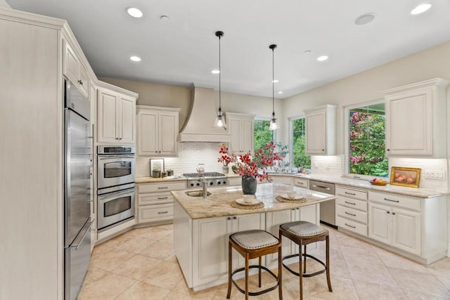 kitchen with appliances with stainless steel finishes, an island with sink, premium range hood, light stone counters, and white cabinetry