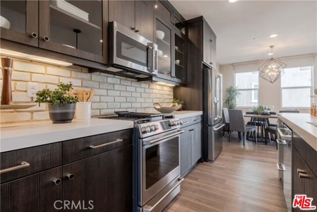 kitchen with appliances with stainless steel finishes, hanging light fixtures, light hardwood / wood-style floors, dark brown cabinetry, and backsplash